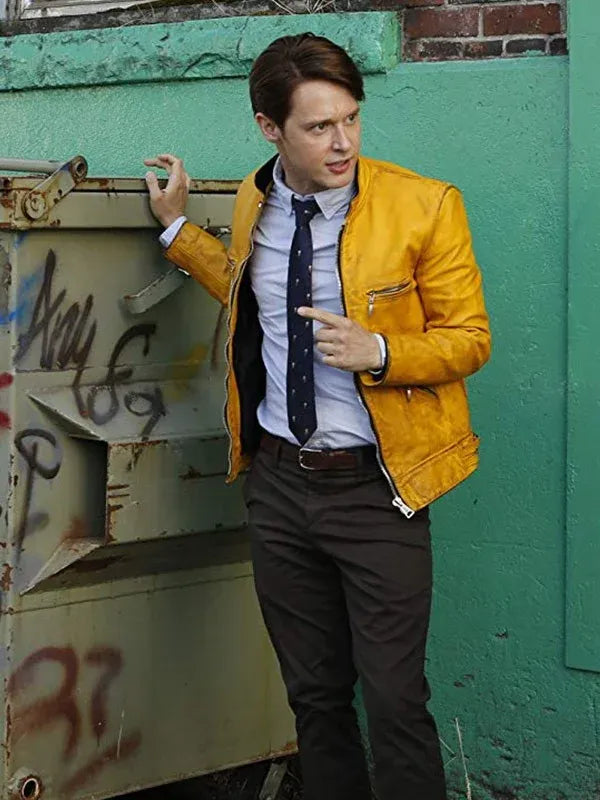 Man in a yellow jacket and dark pants standing against a green wall with graffiti.