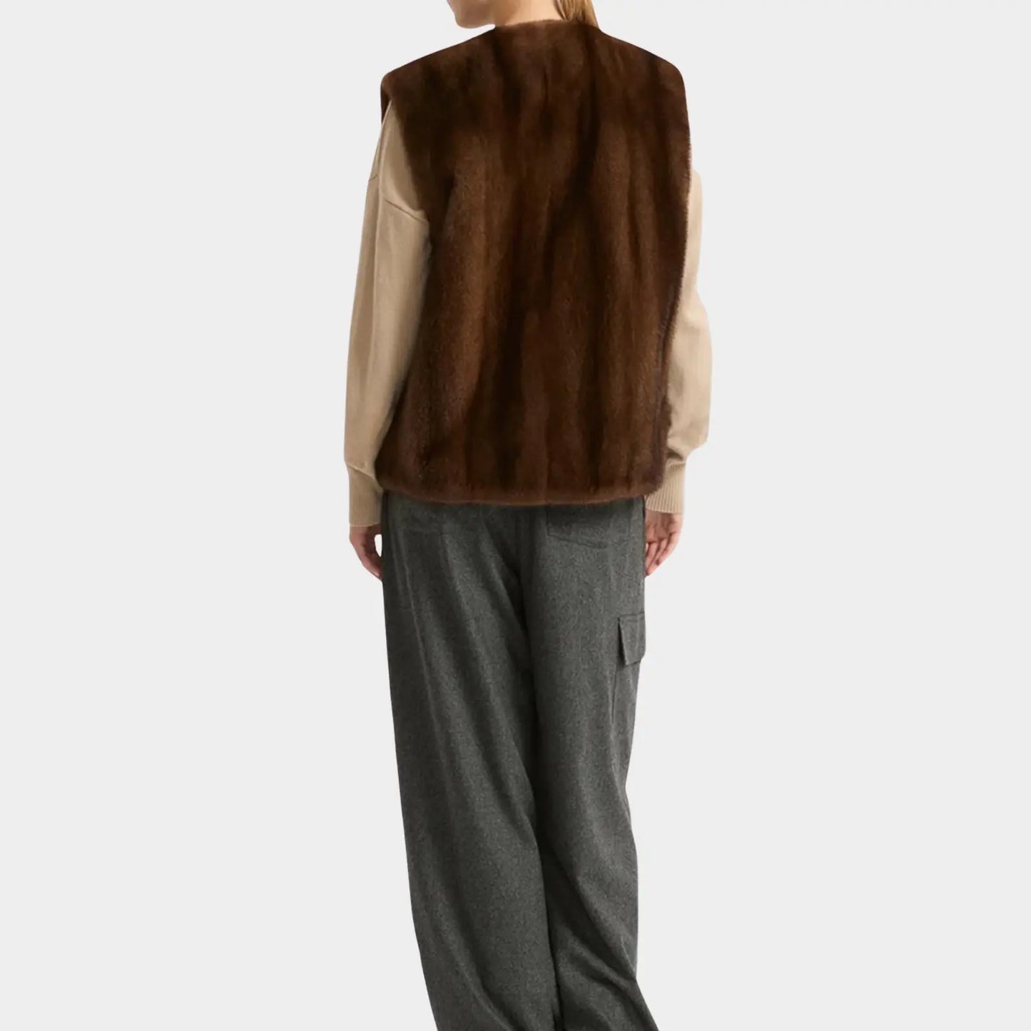 Person wearing a brown fur vest over a beige shirt with gray pants on a white background