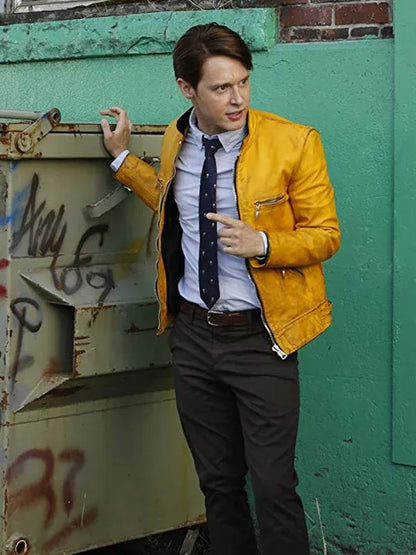 Man in a yellow jacket and dark pants standing against a green wall with graffiti.